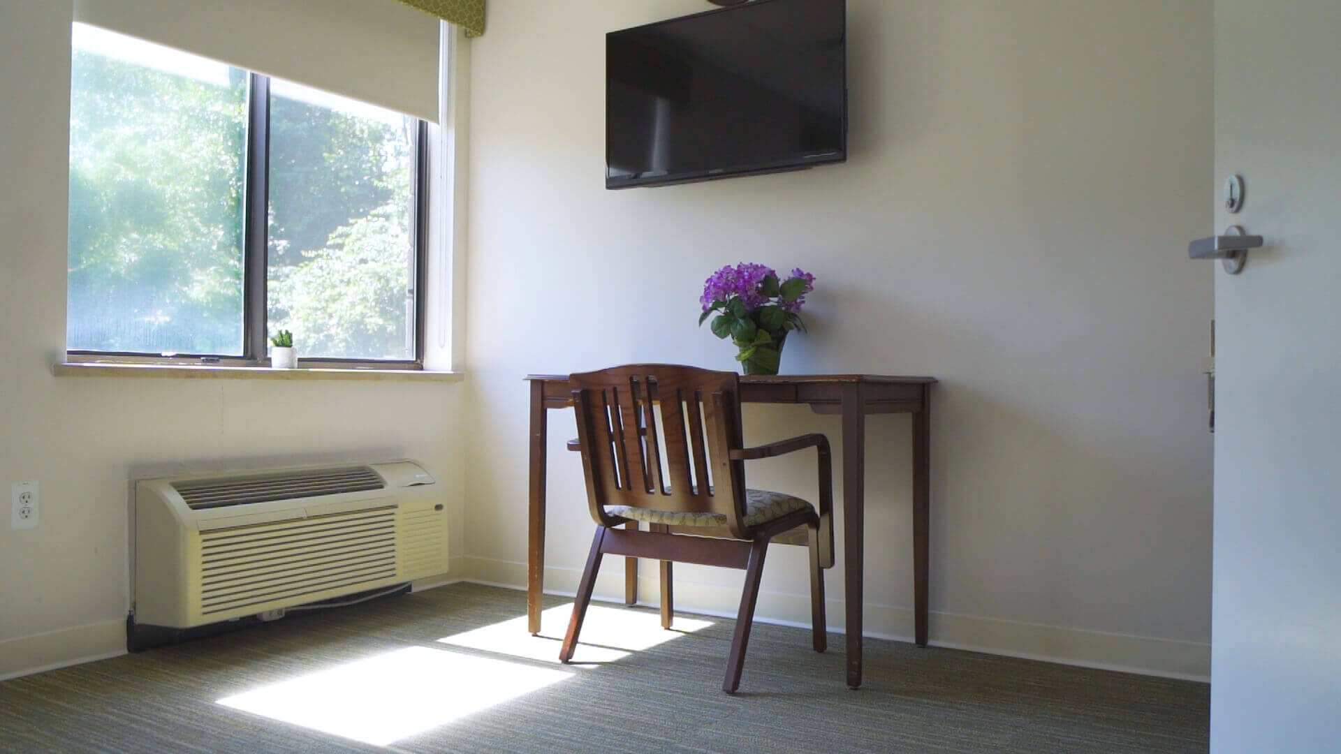 Table with flower vase and chair. Television on the wall and air-condition and one window.