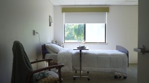 Bed with white pillow and blanket with bed table. Sofa chair on the side with one window on the background.